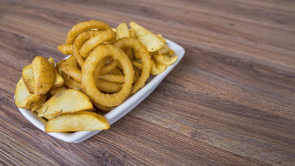 Fotografia tirada decima com uma mesa de fundo. Na esquerda tem um pote de cerâmica branco cheio de batata rústica e anéis de cebola fritos.