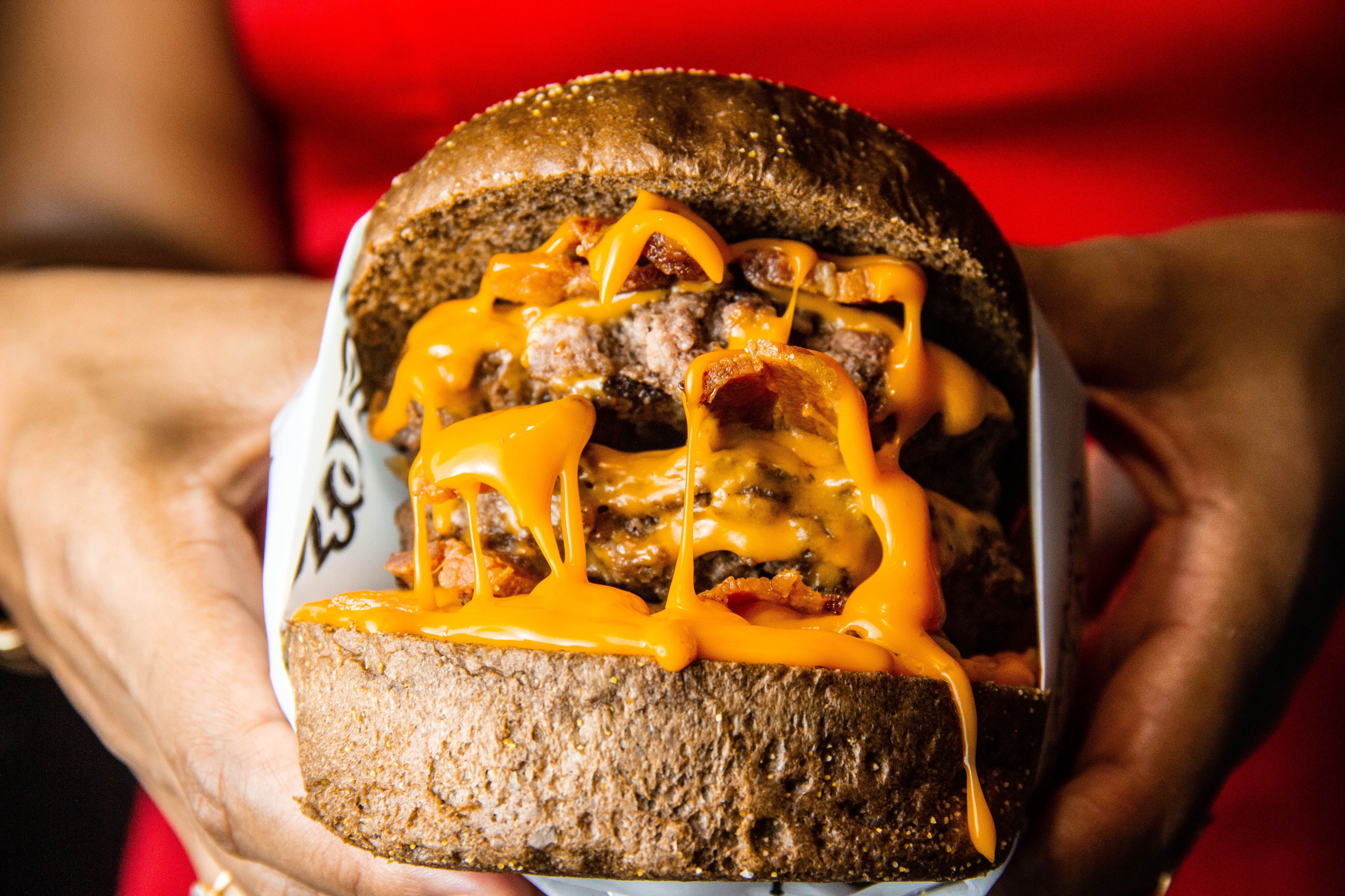 Fotografia de um hambúrguer sendo segurado pelas mãos de uma pessoa de pele morena. O lanche, centralizado na foto, é grande, tem pão escuro, duas carnes e muito molho cheddar. 