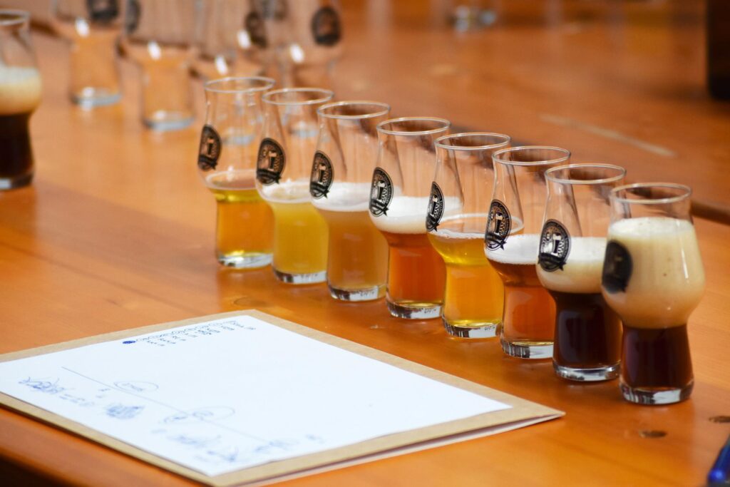 Imagem ilustrativa de oito copos de cerveja, de cores diferentes, enfileirados em cima de uma mesa de madeira. Na frente já um papel com anotações. Imagem inserida no contexto como harmonizar cerveja com hambúrguer