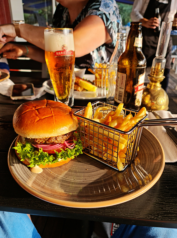 Fotografia ilustrativa de um hambúrguer tradicional à esquerda e uma porção de batata frita à direta. No fundo, aparece um copo e uma garrafa de cerveja e duas pessoas desfocadas. Imagem no contexto como harmonizar cerveja com hambúrguer